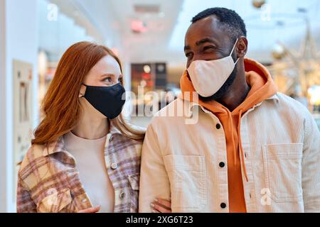 two cheerful smiling lovers in black masks walking, having great time together indoors, holding hands, hugging arm. family, love, relationship Stock Photo