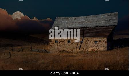 Full moon rising above clouds on the horizon in hilly country over an old abandoned barn shortly after the sun has set. Stock Photo