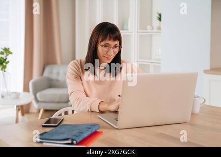 Smiling young woman wearing glasses working remotely, typing on laptop, looking at computer screen, sitting at desk in her home office. Female enjoying elearning process, studying online at home Stock Photo