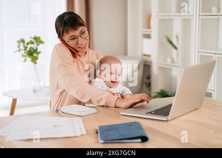 Single mother taking a phone call while holding her baby and using laptop. Happy multitasking business mom making plans with her clients while working from home. New mom balancing motherhood and work. Stock Photo
