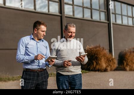 Two men, one wearing a blue shirt and plaid pants and the other wearing a gray sweater and blue jeans, walk and discuss work outside of an industrial Stock Photo