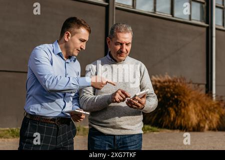 Two men stand outside a building on a sunny day. The younger man is pointing at his phone as the older man looks at the screen. The younger man is wea Stock Photo
