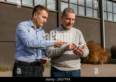 Two men, one younger and one older, are standing in front of a building. The younger man is wearing a light blue button-up shirt and plaid pants, whil Stock Photo