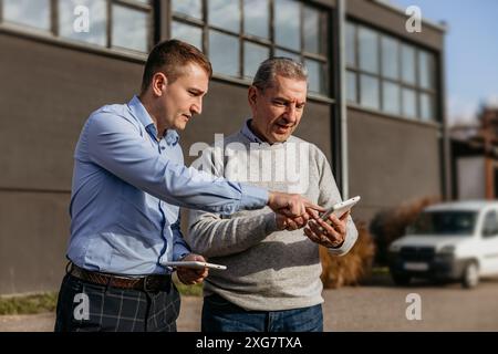 Two men are standing outside a building on a sunny day. A younger man in a blue shirt and plaid pants is pointing at a tablet in the older man's hand. Stock Photo