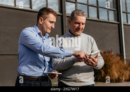 Two men stand outside of a building, one pointing at a tablet screen held by the other, reviewing plans together. Stock Photo