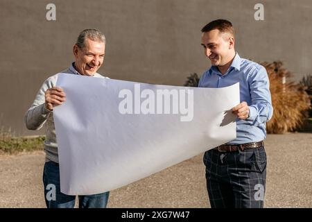 Two men, one older and one younger, stand outdoors, smiling as they hold a large sheet of paper between them. The man on the left is wearing a gray sw Stock Photo