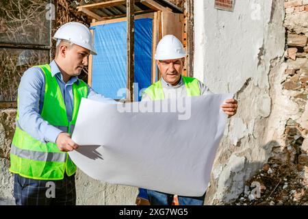 Two construction workers, wearing white hard hats and green safety vests, stand in front of a partially demolished building and examine blueprints. Th Stock Photo