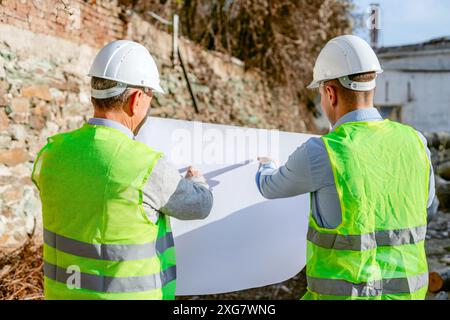 Two construction workers, wearing hard hats and safety vests, stand on a construction site, reviewing plans. Stock Photo