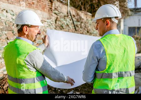 Two construction workers wearing hard hats and safety vests are standing in front of a brick wall at a construction site. Stock Photo