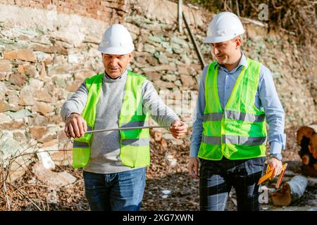 Two construction workers, wearing hard hats and safety vests, stand on a construction site. One worker holds a steel rod while the other observes. The Stock Photo