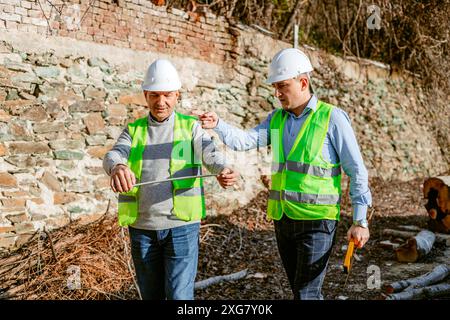 Two construction workers, wearing white hard hats and bright green safety vests, are standing in front of a rock wall. Stock Photo