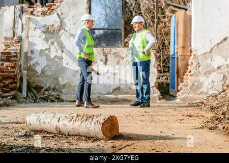 Two construction workers wearing hard hats and bright green vests are standing in front of a demolished building, talking to each other. Stock Photo