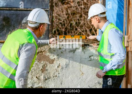 Two construction workers, both wearing white hard hats and neon green safety vests, stand next to a concrete wall on a bright, sunny day. Stock Photo