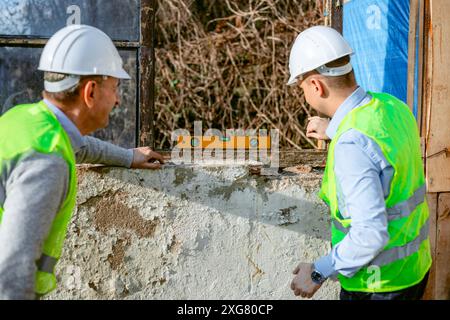 Two construction workers, wearing hard hats and safety vests, carefully check the level of a concrete wall using a yellow level. Stock Photo