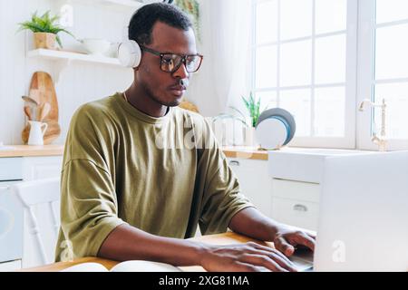 Focused African American man wearing headphones and glasses working on laptop in bright kitchen, studying or remote working. Black guy is freelancer working on online project. Stock Photo