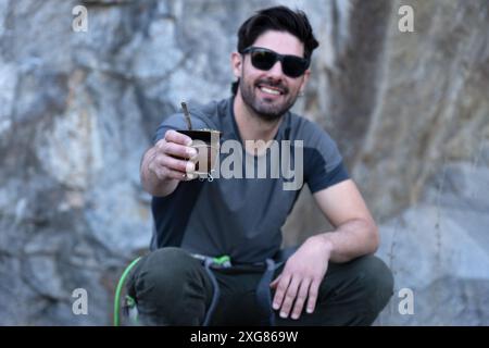 Smiling Latin man in sunglasses and hiking gear, enjoys the outdoors taking a break offering a traditional mate tea. Stock Photo