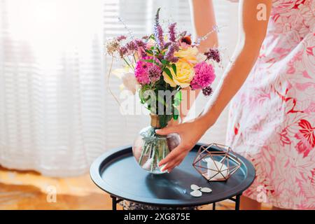 Woman puts bouquet of summer flowers in vase on coffee table at home. Delivery of fresh seasonal flower arrangements for interior decor. Close up Stock Photo