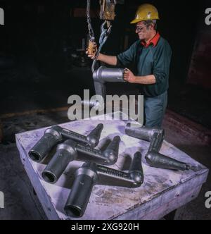 Industrial worker in hard hat operates overhead hoist lifting heavy metal pipe fittings on workshop floor, showcasing manufacturing and rigging proces Stock Photo