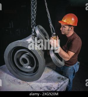 Industrial worker in hard hat operates overhead hoist lifting heavy metal pipe fittings on workshop floor, showcasing manufacturing and rigging proces Stock Photo