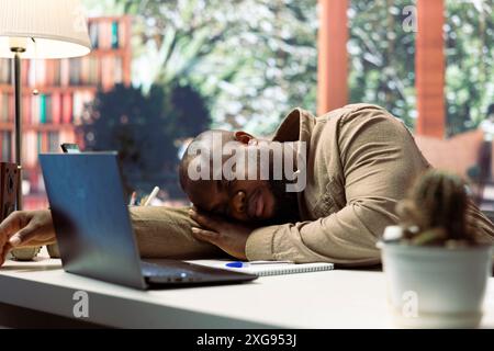 Successful freelancer falling asleep at his home desk setup while he works on laptop, taking a nap after dealing with burnout from online business management. Tired entrepreneur under pressure. Stock Photo
