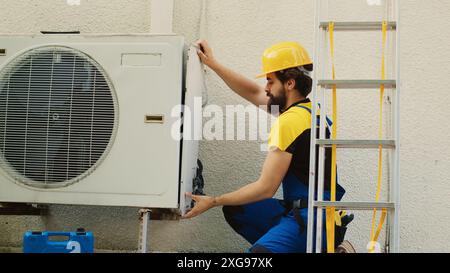 Experienced wireman dissasembling external air conditioner metal side coil panel to check for bad wiring. Meticulous electrician doing repairing on malfunctioning hvac system Stock Photo