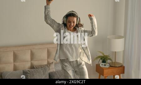 Cheerful woman jumping dancing on bed bedroom home mobile phone enjoy joyful headphones listening music female technology singing happy excited energy Stock Photo