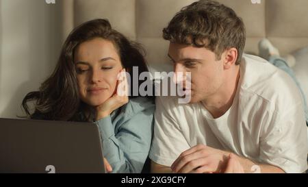 Man woman boyfriend girlfriend female male lying on bed bedroom laptop watching movie cinema couple online computer tech cyberspace girl guy together Stock Photo