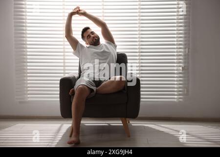 Man sitting on armchair near window blinds indoors Stock Photo