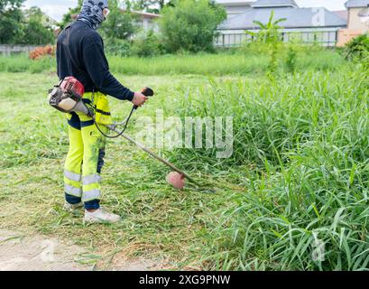 Worker mowing tall grass with petrol lawn trimmer backyard. The gardener man mows the grass with a hand lawn mower. Stock Photo