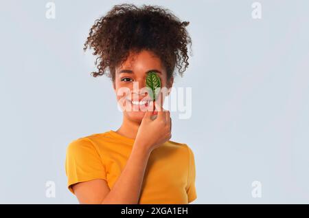 Natural beauty. Happy Afro american girl in yellow tshirt with curly hair covering one eye with green leaf, broadly smiling while looking directly Stock Photo