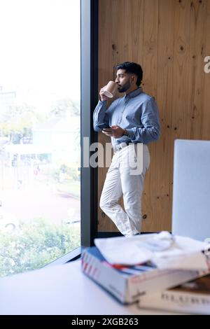 Drinking coffee and using smartphone, businessman standing by office window Stock Photo