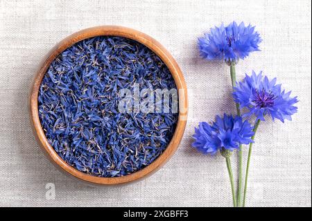 Dried blue cornflower petals in wooden bowl on linen fabric. Edible flowers of Centaurea cyanus, also known as bachelors button. Stock Photo