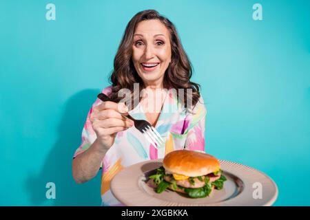 Photo portrait of attractive senior woman eating burger dressed stylish colorful clothes isolated on cyan color background Stock Photo