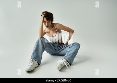 Young woman in jeans and white tank top sits on the floor in a stylish pose. Stock Photo