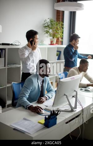 African american man working on his personal computer, surrounded by his colleagues in office.Handsome young man at his desk. Stock Photo