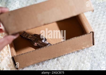 A person opening a cardboard box containing a wristwatch with a metal band, placed on bubble wrap. Stock Photo