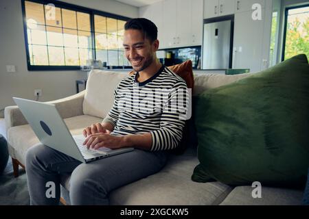 Man smiles while leisurely using laptop computer on couch by window in house Stock Photo