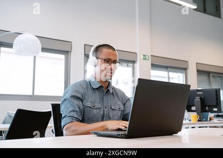 A man wearing headphones, focused on his laptop in a contemporary office setting with large windows. Stock Photo