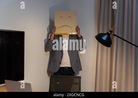 Portrait of a businessman covering his face with a paper mask with a sad face, drawn on it Bad negative depression from the anonymous person in the co Stock Photo