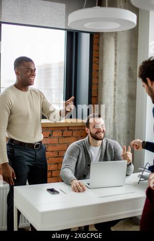 Couple of multiracial male software developers sharing ideas with their caucasian boss working on laptop in modern spacious office with loft interior Stock Photo