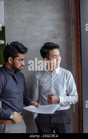 Mix raced couple of male business partners analyzing reports against grey wall. Indian and Chinese men in formal shirts standing in grey office boardr Stock Photo