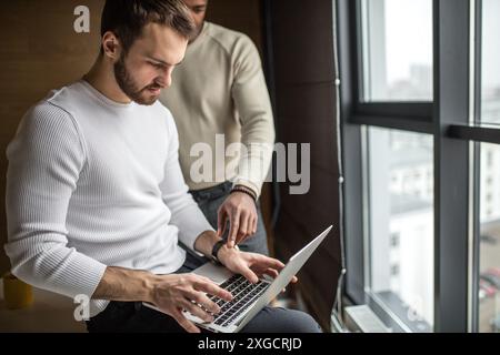 Two concentrated black and white businessmen in casual wear discussing project sitting at office desk, near the window, using pc laptop, looking at sc Stock Photo