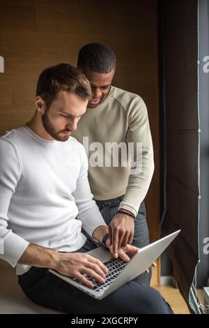 Two concentrated black and white businessmen in casual wear discussing project sitting at office desk, near the window, using pc laptop, looking at sc Stock Photo