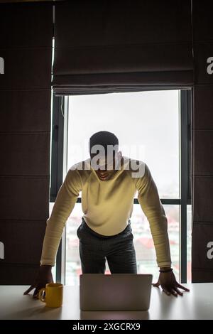 Concentrated African man in white sweater looking at laptop while standing in dark shades room with window day light . Stock Photo