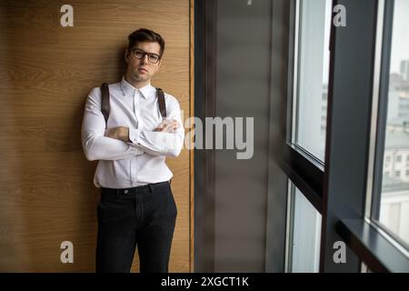 Portrait of stylish young Caucasian officer wearing spectacles, dressed in white and black formal clothes, posing isolated with folded hands against c Stock Photo