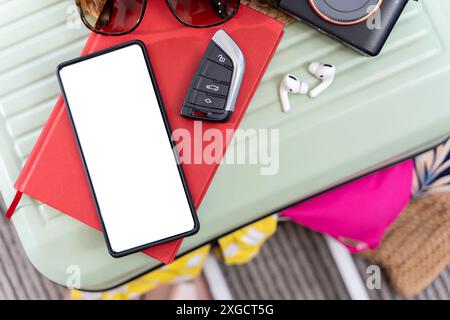 A green suitcase sits on a table. On top of the suitcase is a cell phone, car keys, sunglasses, earbuds, and a camera. The earbuds are white and have Stock Photo