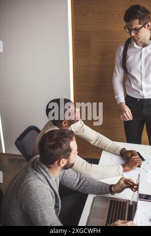 Positive young multiracial male team sitting at conference meeting table listening to speaker presenting new project in office, Business Team Meeting Stock Photo