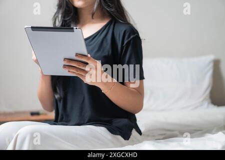 A woman sitting on a bed using a tablet. She is wearing a black t-shirt and white pants. The background is minimalistic with white bedding. Stock Photo