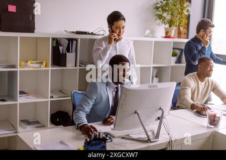 Two african american businessmen working on pc while their colleagues speaking on smartphone standing behind Stock Photo