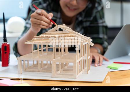 Young elegant female architect with paper and pen bending over model of new house and yard and pointing at roof while working over project Stock Photo